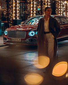 a man standing next to a red car in front of a building with christmas lights