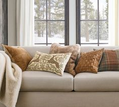 a couch with pillows and blankets on it in front of two large windows that look out onto the snowy woods