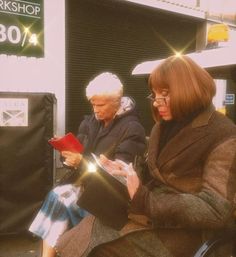 two women are sitting on a bench looking at their cell phones while one woman is holding a red folder