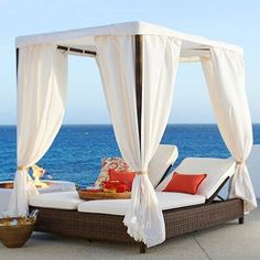 an outdoor bed with white drapes and pillows on the floor next to the ocean