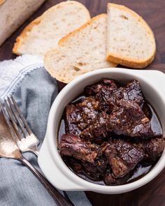 a white bowl filled with meat and bread on top of a wooden table next to a fork