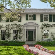 a large white house with green shutters and flowers on the front lawn in front of it