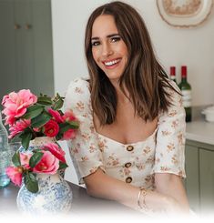 a woman sitting at a table with flowers in front of her and a bottle of wine behind her