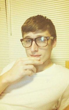 a young man wearing glasses sitting in front of a window with his hand on his chin