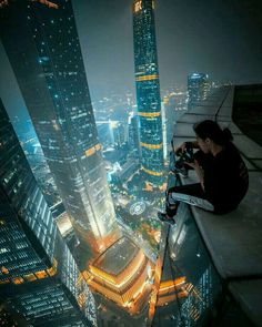 a man sitting on top of a tall building looking at the city lights in the distance