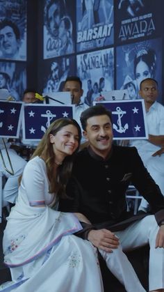 a man and woman sitting next to each other in front of a wall with pictures on it