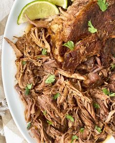 a white plate topped with meat and garnished with cilantro, lime wedges