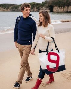 a man and woman are walking on the beach with a tote bag in hand