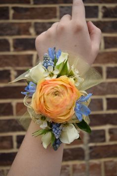 a wrist corsage with an orange rose, blue flowers and greenery is shown in front of a brick wall