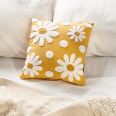 a yellow pillow with white daisies on it sitting on a bed next to a blanket