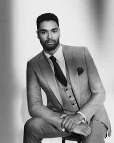 a black and white photo of a man in a suit sitting on a stool wearing a tie