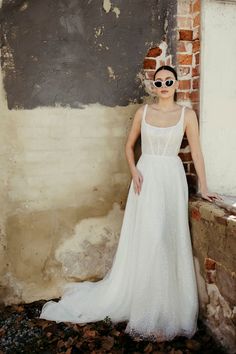 a woman in a white dress standing next to a brick wall and wearing sunglasses on her head