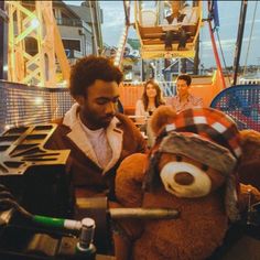 a man sitting next to a brown teddy bear on top of a roller coaster at an amusement park