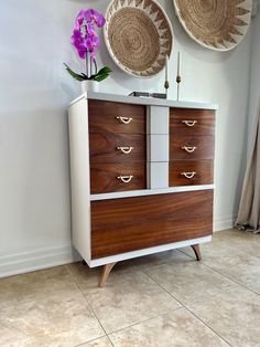 a white and brown dresser with two baskets on it's wall next to a purple flower