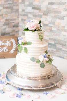 a white wedding cake with pink and blue flowers on top is sitting on a silver platter