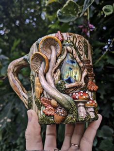 a person holding up a ceramic mug with mushrooms and leaves on the outside, in front of some trees