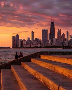 two people are sitting on steps overlooking the water and skyscrapers in the background at sunset