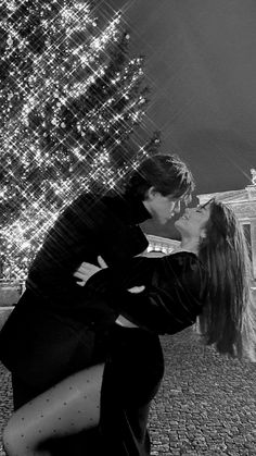 a man and woman kissing in front of a christmas tree with lights on the building behind them