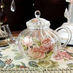 a glass teapot with pink roses on it sitting on top of a floral table cloth