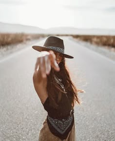 a woman with long hair wearing a black hat pointing at the camera on an empty road