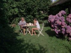 two women sitting at a table outside in the grass with purple hydrangeas behind them