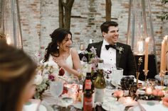 a bride and groom sitting at a table with candles in front of them, laughing