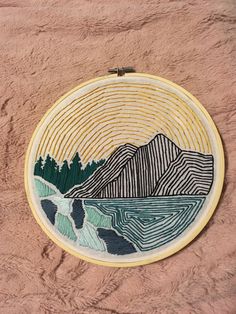 a hand embroidered wall hanging with mountains and trees in the background on a pink surface