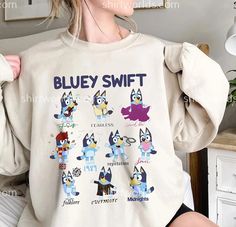 a woman sitting on a bed wearing a bluey swift sweatshirt with cartoon characters printed on it