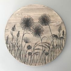 a round wooden plaque with dandelions and grasses painted on the front in black ink