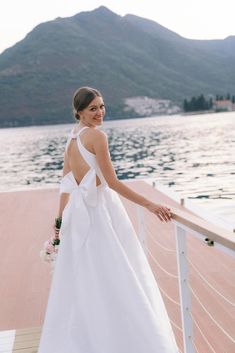 a woman in a white dress standing on a dock next to the water and mountains