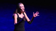 a woman in a black dress holding her hands out to the side while standing on stage