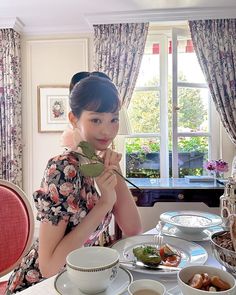 a woman sitting at a table with plates and cups on it, in front of a window