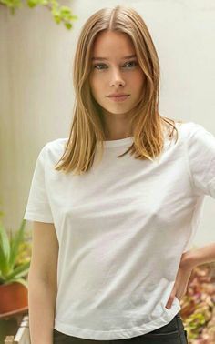 a woman in white shirt and black pants standing next to potted plants with her hand on her hip