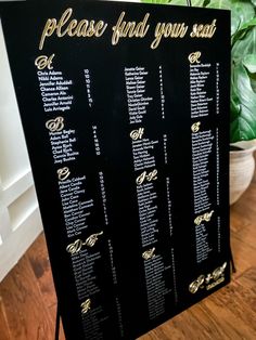 a black and gold wedding seating chart on a wooden table next to a potted plant