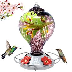 a hummingbird is flying near a glass vase with flowers on it and another bird in the background