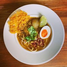 a white plate topped with soup and noodles next to an egg on top of a wooden table