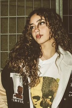 a woman with long curly hair wearing a t - shirt and jacket standing in front of a cage