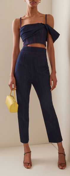 a woman is standing in front of a white wall holding a yellow purse and wearing blue pants