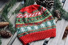 a red and green knitted hat sitting on top of a wooden table next to pine cones