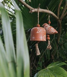 two bells hanging from a tree branch in front of some plants and trees with green leaves