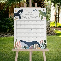 a baby shower calendar sitting on top of a wooden easel in front of a palm tree