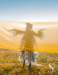 a woman is standing in a field with her arms spread out to the side, holding a yellow cloth over her head