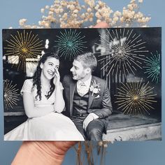 a person holding up a card with fireworks in the background and a couple sitting on a bench