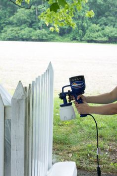 a woman using a paint sprayer on a white picket fence in front of a field