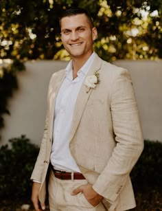 a man in a tan suit and white shirt smiles at the camera with his hands in his pockets