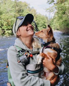 a woman holding a dog in her arms while standing next to a river with trees