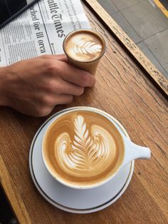 a person holding a cup of coffee on top of a wooden table next to a newspaper