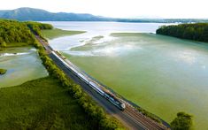 an aerial view of a train on the tracks next to a body of water with mountains in the background