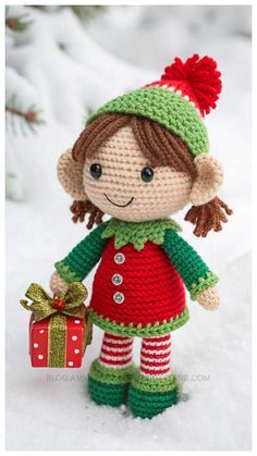 a crocheted doll is holding a gift box in the snow, wearing a green and red outfit