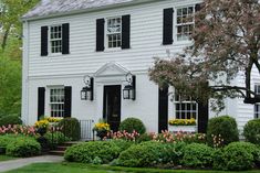 a white house with black shutters and flowers in the front yard on a sunny day